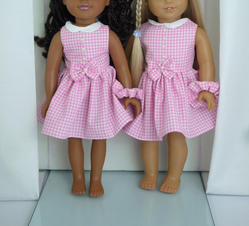 May include: Two dolls dressed in matching pink and white gingham dresses. The dresses feature white collars, pearl buttons, and a bow at the waist. Each doll also has a matching ruffled cuff. One doll has dark hair, and the other has blonde hair.