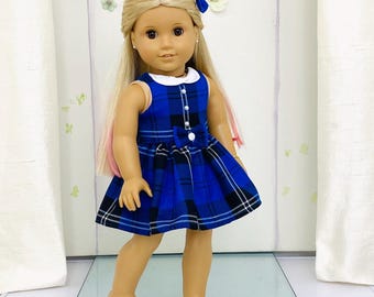 Op de afbeelding: Een pop met een blauw geruite jurk met een witte kraag en een bijpassende blauwe strik in haar blonde haar. De jurk heeft een geplooide rok en is aan de voorkant dichtgeknoopt. De pop staat op een doorzichtig oppervlak.