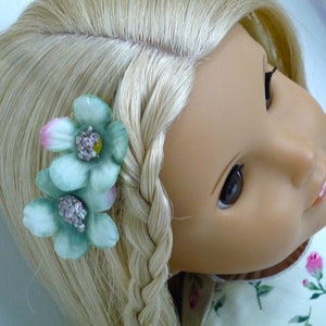 May include: A doll with blonde hair styled in a plait, decorated with two light green flower hair clips. The flowers have pink and yellow accents. The doll is wearing a floral print dress.