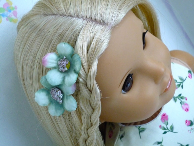 May include: A doll with blonde hair styled in a plait, decorated with two light green flower hair clips. The flowers have pink and yellow accents. The doll is wearing a floral print dress.