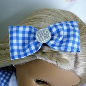 May include: A blue and white gingham bow hair accessory with a rhinestone centre. The bow is attached to a doll's blonde hair, with a matching gingham dress visible. This accessory adds a touch of charm.