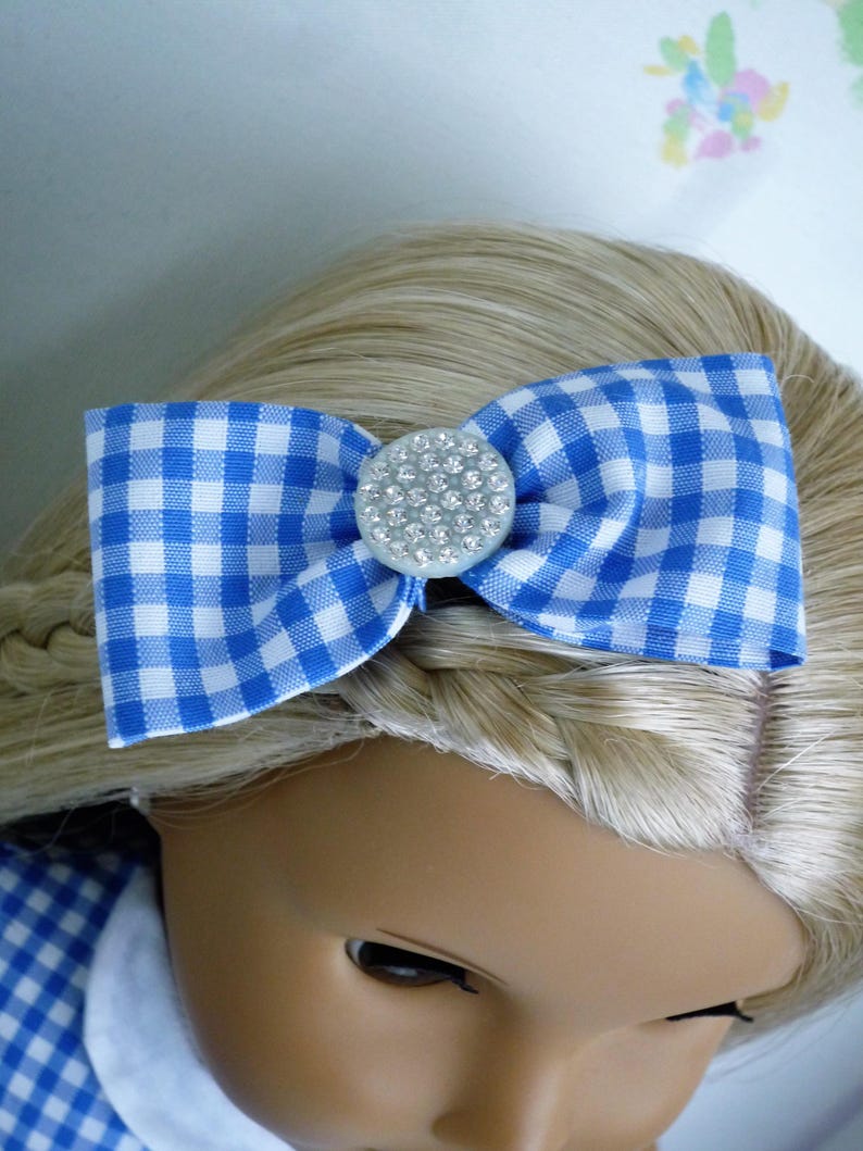 May include: A blue and white gingham bow hair accessory with a rhinestone centre. The bow is attached to a doll's blonde hair, with a matching gingham dress visible. This accessory adds a touch of charm.