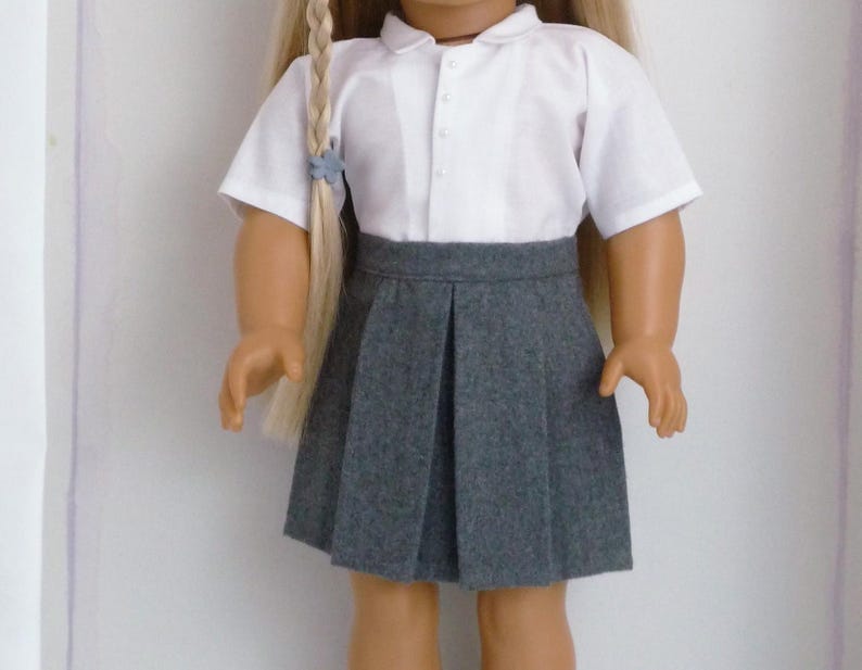 May include: A doll wearing a school uniform consisting of a white short-sleeved collared shirt and a gray pleated skirt. The doll has long blonde hair styled in a braid.