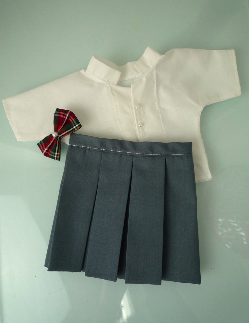 May include: A doll-sized school uniform set, featuring a white short-sleeved shirt with pearl buttons, a pleated gray skirt, and a plaid bow tie with a pearl center. The outfit is designed for a doll.