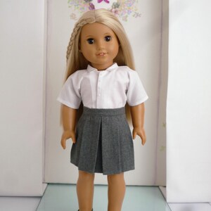 May include: A doll with long blonde hair wearing a white collared shirt, a gray pleated skirt, and black shoes. The doll is standing in front of a wall with a floral wreath and the letter "K".