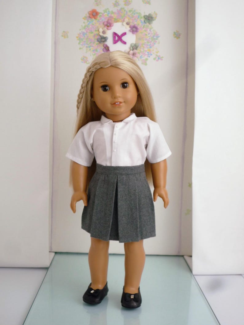May include: A doll with long blonde hair wearing a white collared shirt, a gray pleated skirt, and black shoes. The doll is standing in front of a wall with a floral wreath and the letter "K".