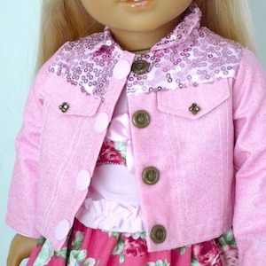 May include: A pink doll jacket with a sequined collar and decorative buttons. The jacket has two pockets and is worn over a floral dress. The buttons are a bronze color. The jacket is made of a soft material.