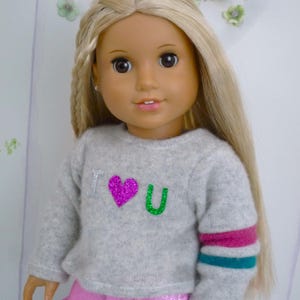 Könnte beinhalten: Eine Puppe mit langen blonden Haaren, die einen grauen Pullover mit "I ♥ U" in Silber, Pink und Grün trägt. Die Puppe trägt auch eine rosa Hose und hat einen Sternanhänger. Der Pullover hat rosa und türkisfarbene Streifen am Ärmel.