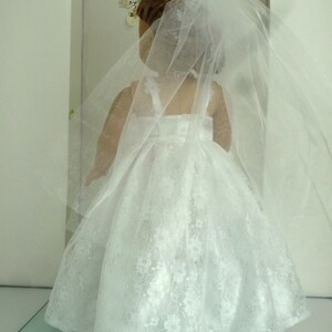 May include: A white lace wedding dress with a long veil for a doll. The dress has a sweetheart neckline and a fitted bodice. The veil is made of tulle and has a scalloped edge.