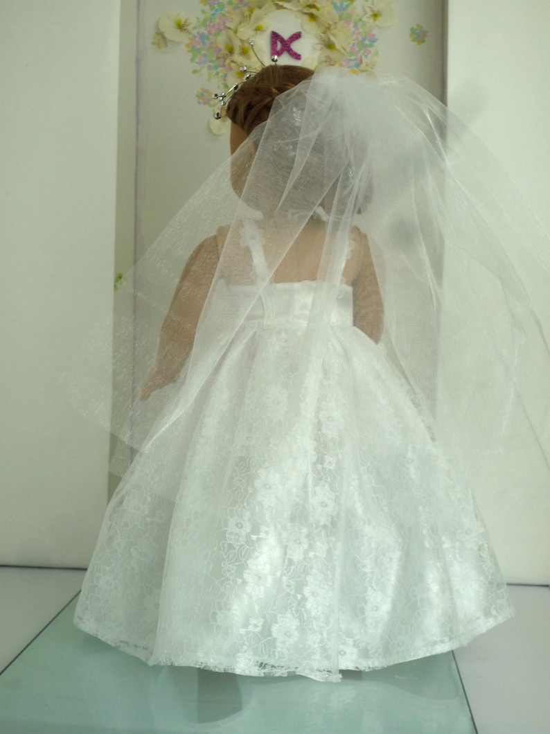 May include: A white lace wedding dress with a long veil for a doll. The dress has a sweetheart neckline and a fitted bodice. The veil is made of tulle and has a scalloped edge.