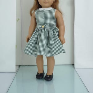 May include: A green and white gingham dress with a white collar and a yellow button. The dress is worn by a doll with brown hair and black shoes.