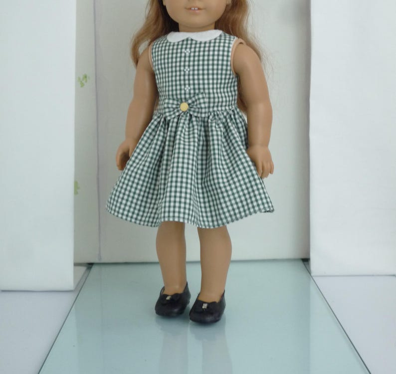 May include: A green and white gingham dress with a white collar and a yellow button. The dress is worn by a doll with brown hair and black shoes.
