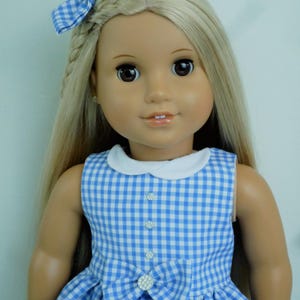 May include: A doll with long blonde hair and brown eyes, dressed in a blue and white gingham dress with a white collar and a bow. A matching gingham bow with a rhinestone accent is in the doll's hair. The dress has a bow with a rhinestone centre.