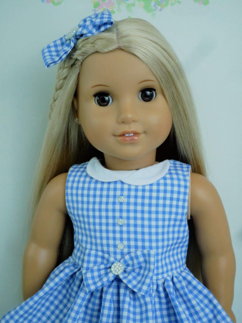 May include: A doll with long blonde hair and brown eyes, dressed in a blue and white gingham dress with a white collar and a bow. A matching gingham bow with a rhinestone accent is in the doll's hair. The dress has a bow with a rhinestone centre.