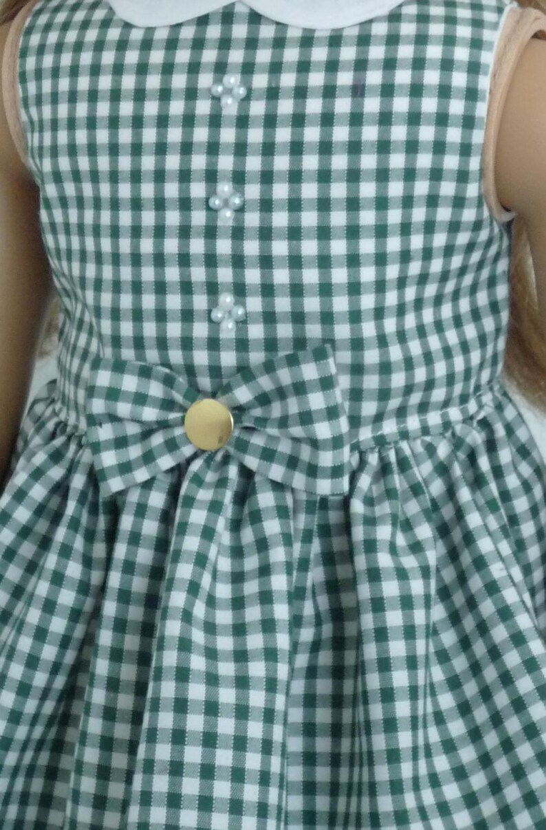 May include: A green and white gingham dress with a white collar and a bow at the waist. The dress has three pearl-like buttons down the front.