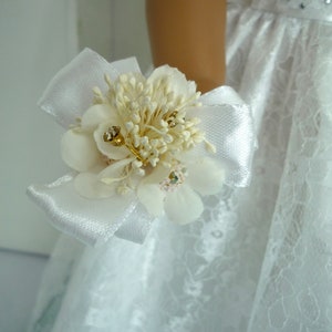 May include: A white satin ribbon with a small bouquet of white flowers and gold accents. The bouquet is attached to a white lace dress.