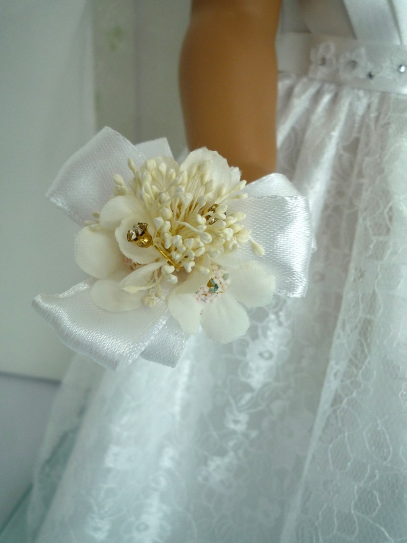 May include: A white satin ribbon with a small bouquet of white flowers and gold accents. The bouquet is attached to a white lace dress.