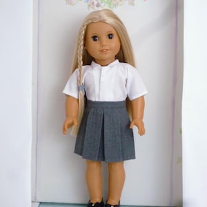 May include: A doll wearing a white short-sleeved shirt, a gray pleated skirt, and black shoes. The doll has long blonde hair styled in a braid. A floral wreath with the letter "K" is in the background.