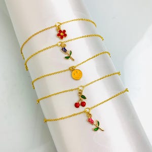 Peut inclure: Bracelets à chaîne dorée avec des breloques en émail coloré. Les breloques comprennent une fleur rouge, une rose bleue, un smiley jaune, des cerises rouges et une rose rose. Les bracelets sont présentés sur une surface blanche.