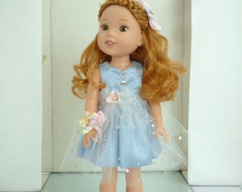 May include: A doll with red hair wearing a light blue dress with a tulle overlay. The doll has a flower bracelet and matching shoes. A flower crown and a heart pendant necklace complete the outfit. The doll is standing in a display case.
