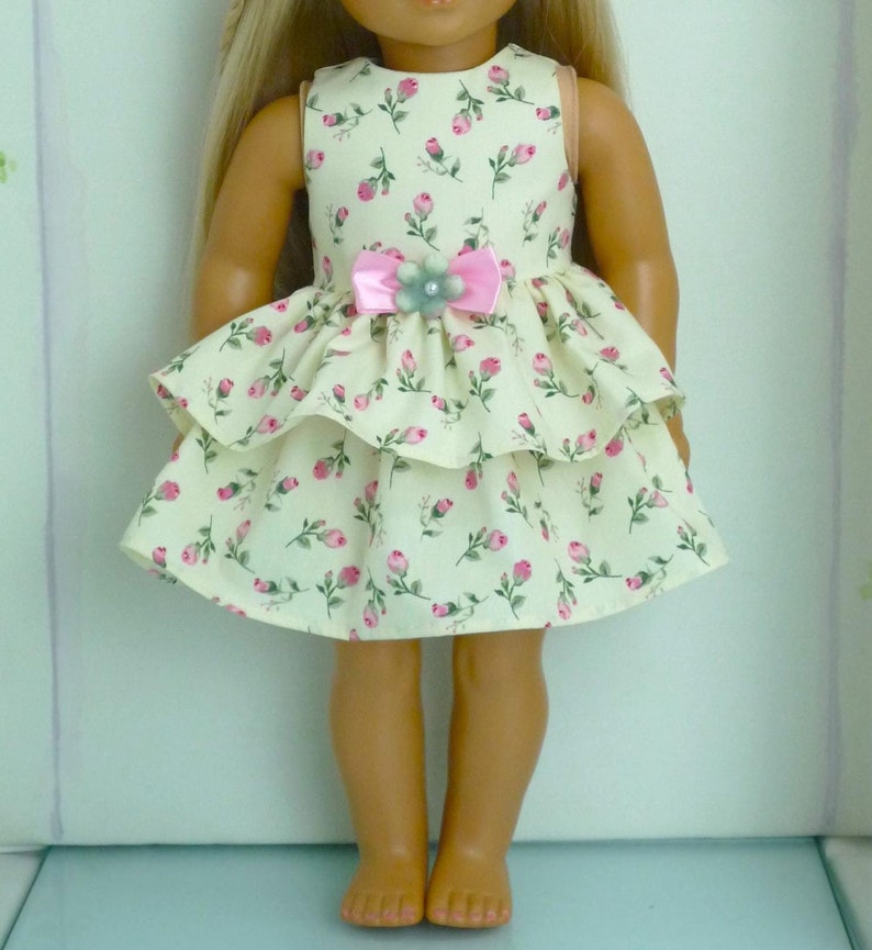 May include: A doll's dress in cream fabric, patterned with pink roses. It has a tiered skirt and a pink bow with a green flower detail at the waist. The sleeveless top and skirt are clearly visible.