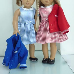 May include: Two dolls wearing gingham dresses and matching jackets. One doll is wearing a blue gingham dress with a white collar and a blue jacket. The other doll is wearing a red gingham dress with a white collar and a red jacket.