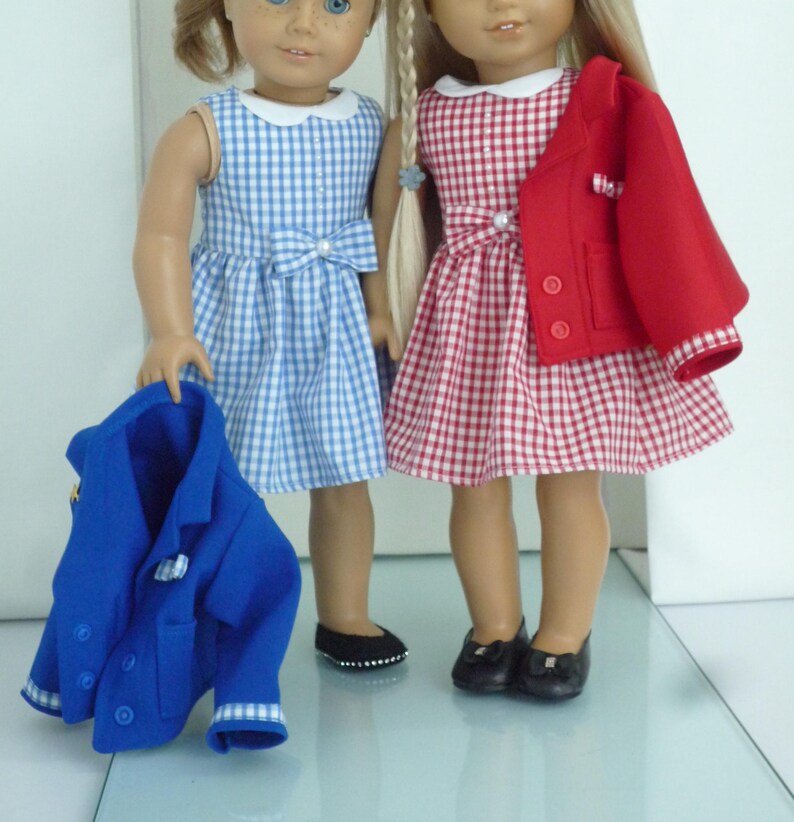 May include: Two dolls wearing gingham dresses and matching jackets. One doll is wearing a blue gingham dress with a white collar and a blue jacket. The other doll is wearing a red gingham dress with a white collar and a red jacket.