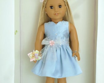 May include: A doll wearing a light blue dress with a white bow and a floral accent at the waist. The doll has long blonde hair and is holding a small bouquet. The doll is also wearing flower-adorned sandals.