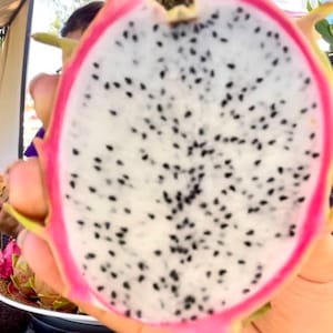 May include: A halved dragon fruit showing a bright pink outer rind and a white interior flesh speckled with numerous small black seeds. The fruit is oval in shape with green-tipped scales visible along the edges of the vibrant pink skin.