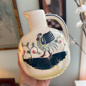 May include: Cream-colored ceramic pitcher with a rooster illustration. The rooster is depicted with blue, yellow, red, and green accents. The pitcher has a dark green base and handle, with pink floral accents.