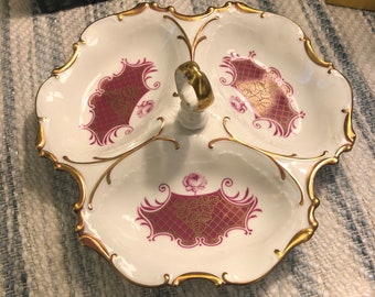 May include: A white porcelain serving dish with three sections, each decorated with a pink and gold floral design. The dish has a gold rim and a gold handle in the center.