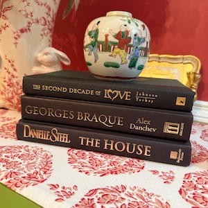 May include: A stack of three hardback books with gold lettering, topped with a small, round, hand-painted vase. The books are titled "The Second Decade of Love", "Georges Braque", and "Danielle Steel The House". The vase features figures in colorful clothing.