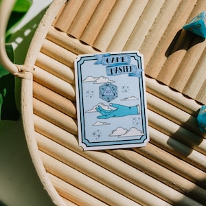 May include: A blue and white sticker with a hand holding a blue 20-sided die. The sticker says "Game Master" in a banner above the die. The sticker is on a wooden tray with blue dice.
