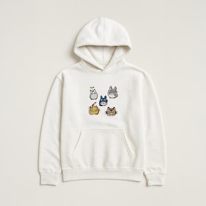 May include: A cream-coloured hooded sweatshirt with a kangaroo pocket and a drawstring hood. The front displays an embroidered design of six cartoon characters in various colours. The characters include a sprout, a blue creature, a grey creature, a flame, and a cat.
