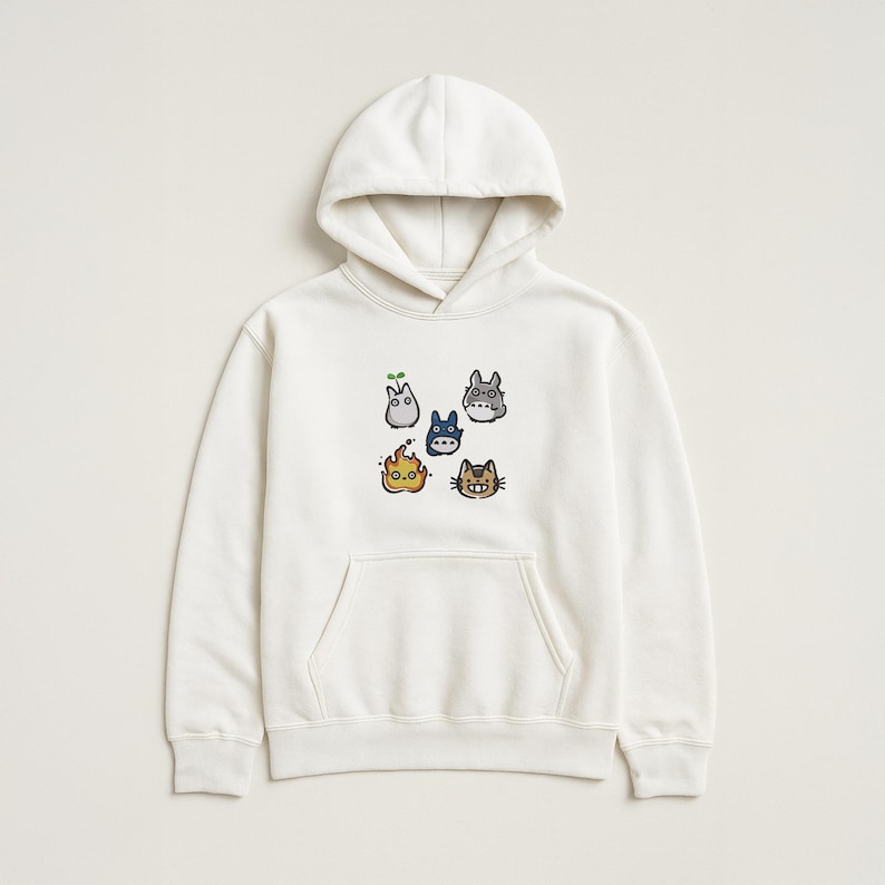 May include: A cream-coloured hooded sweatshirt with a kangaroo pocket and a drawstring hood. The front displays an embroidered design of six cartoon characters in various colours. The characters include a sprout, a blue creature, a grey creature, a flame, and a cat.