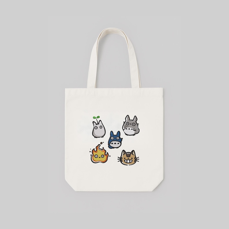 May include: A white tote bag with a design featuring cartoon characters. The bag has a rectangular shape and two handles. The cartoon characters are in various colours and shapes, including a blue creature and a cat-like character.