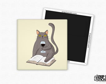 May include: Square magnet featuring a cartoon cat with gray fur, yellow eyes, and a wine glass. The cat sits on an open book, holding the glass of red wine. The background is a light beige color. The magnet is next to a black magnet.