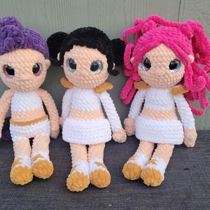 May include: Three handmade crochet dolls with purple, black, and pink hair. Each doll has a light skin tone, large glittery eyes, and wears a white dress with gold accents. The dolls are displayed together.