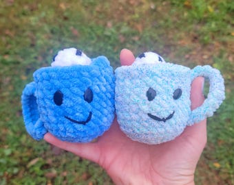 May include: Two crocheted mugs with smiling faces. One mug is blue and the other is light blue with white flecks.