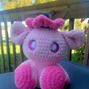 Puede incluir: Un juguete de peluche rosa tejido a crochet con una flor en la cabeza. El juguete tiene ojos brillantes y está sentado en una superficie gris.