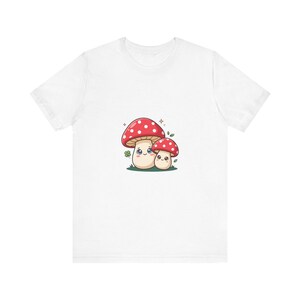 Könnte beinhalten: Ein weißes Kurzarm-T-Shirt mit einer zentralen Grafik von zwei Cartoon-Pilzen mit roten Kappen, weißen Punkten und lächelnden Gesichtern. Der größere Pilz befindet sich links und der kleinere rechts, beide auf einem kleinen Stück grünem Gras.