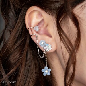 May include: A silver ear cuff with a delicate chain and blue flower charms. The chain dangles from the cuff and ends with a larger blue flower charm.