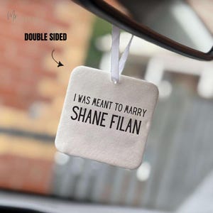 May include: A white, square car air freshener with the text "I WAS MEANT TO MARRY SHANE FILAN" in black. It hangs from a white ribbon and is labeled "DOUBLE SIDED". The background is blurred.
