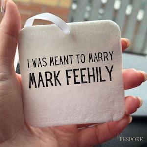 May include: A square, off-white fabric tag with the text "I WAS MEANT TO MARRY MARK FEEHILY" in black. A white ribbon loop is attached at the top. The tag is held in a hand.
