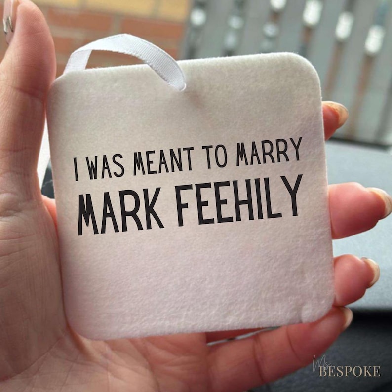 May include: A square, off-white fabric tag with the text "I WAS MEANT TO MARRY MARK FEEHILY" in black. A white ribbon loop is attached at the top. The tag is held in a hand.