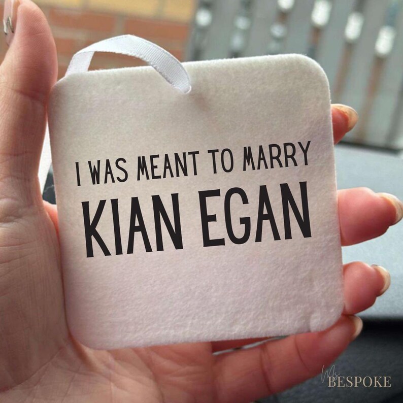 May include: A square, off-white tag with the text "I WAS MEANT TO MARRY KIAN EGAN" in black. A white ribbon loop is attached at the top. The tag is held in a hand.