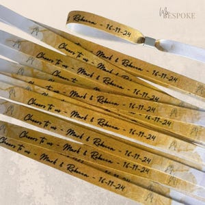 May include: Wedding wristbands in gold and white, featuring the text "Cheers to us - Mark & Rebecca - 16-11-24" in black script. The wristbands have a gold watercolour design and a champagne glass graphic.