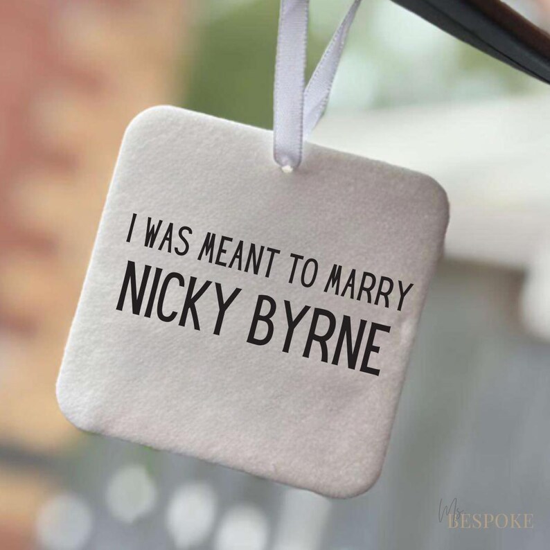 May include: A square, off-white tag with the text "I WAS MEANT TO MARRY NICKY BYRNE" in black. The tag is suspended by a white ribbon. The background is blurred, suggesting an indoor setting.