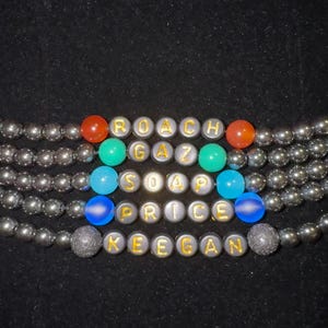May include: Close-up of several beaded bracelets with the words "ROACH GAZ SOAP PRICE KEGAN" spelled out in gold letters. The bracelets are made of gray beads and colorful accent beads against a black background.