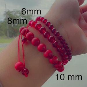 May include: Three red beaded bracelets, with bead sizes of 6mm, 8mm, and 10mm, are displayed on a wrist. The bracelets are made of round, polished beads and are strung on a red cord. The text "6mm", "8mm", and "10 mm" is visible.